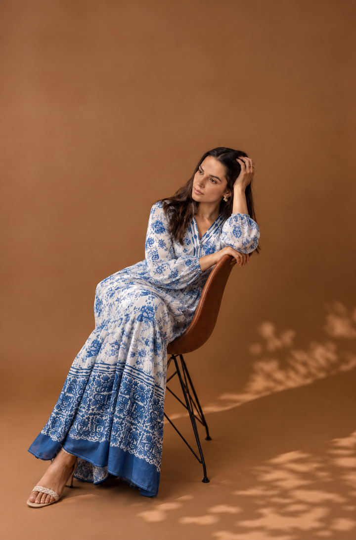 A woman wearing a long-sleeved blue and white patterned maxi dress sits on a brown chair against a solid brown studio backdrop.